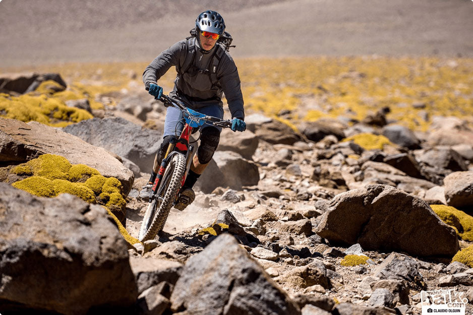 A person riding a bike uphill across rough rocky territory wearing a helmet, sunglasses, and backpack