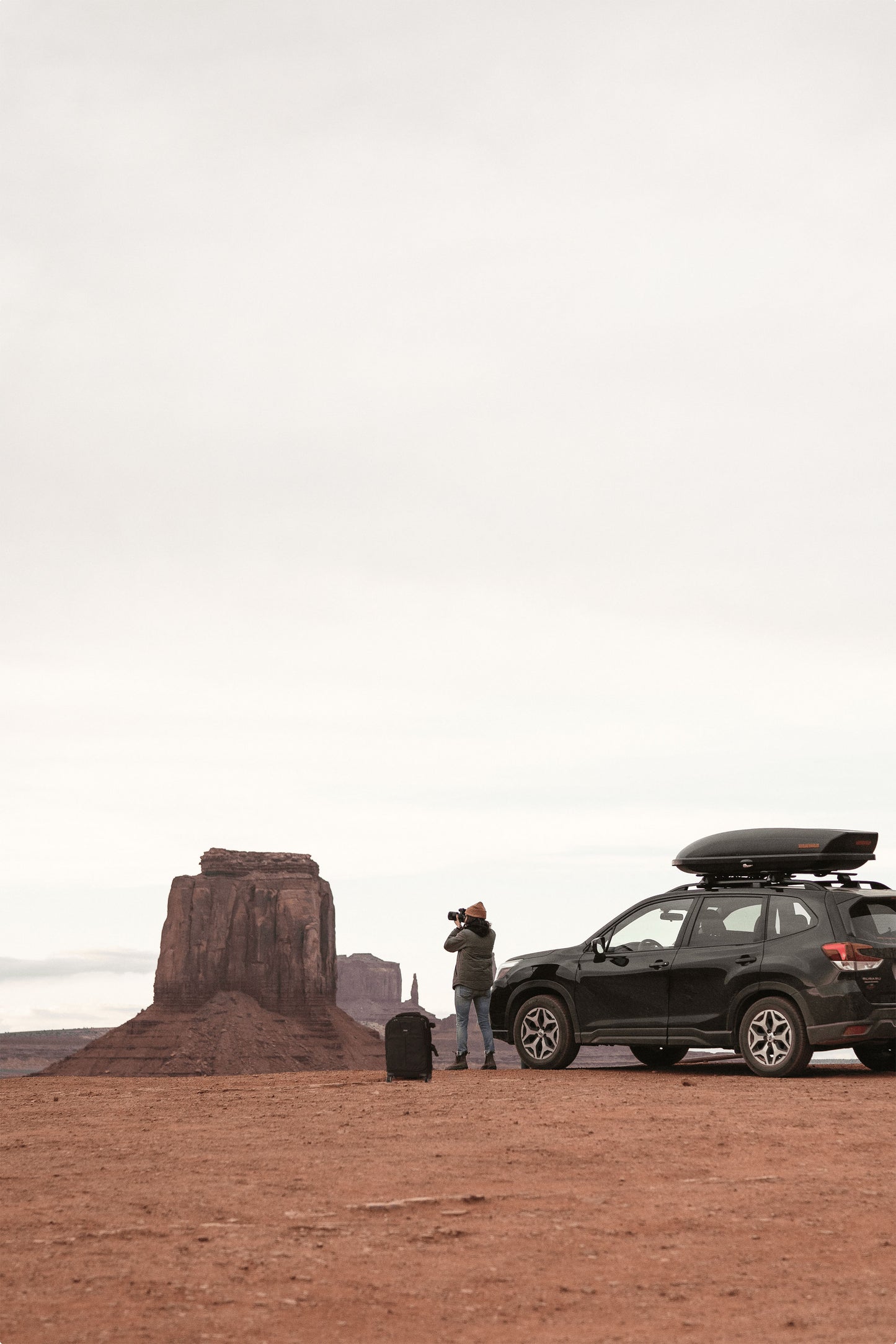 A photographer holding a camera to his face, and standing in-between a roller backpack and a black car