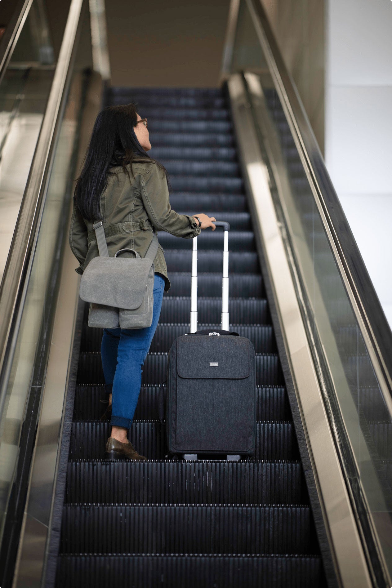 A women riding an escalator up, wearing a small camera bag and holding a rolling luggage