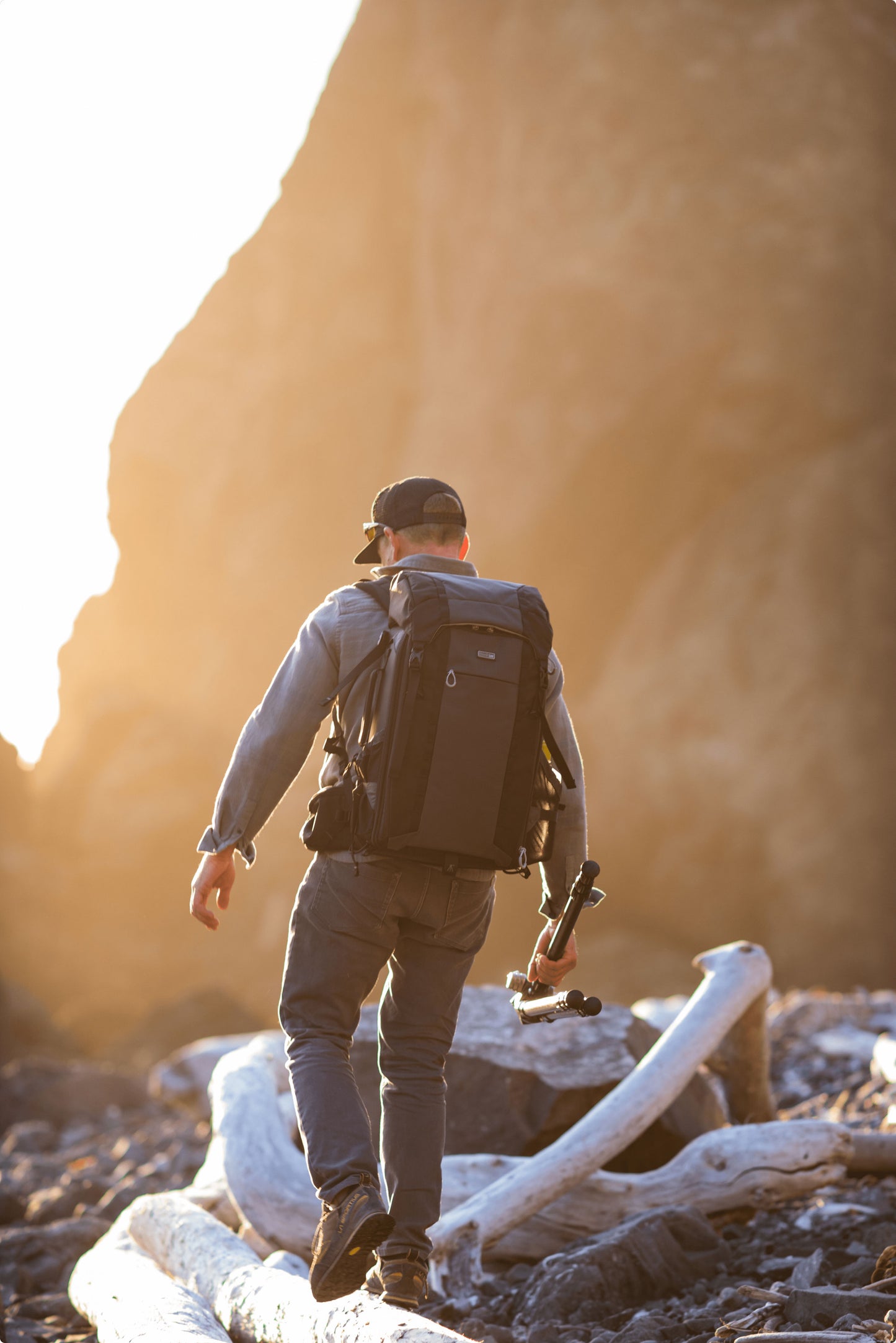 A man balancing on a log during sunrise holding a tripod and wearing a backpack