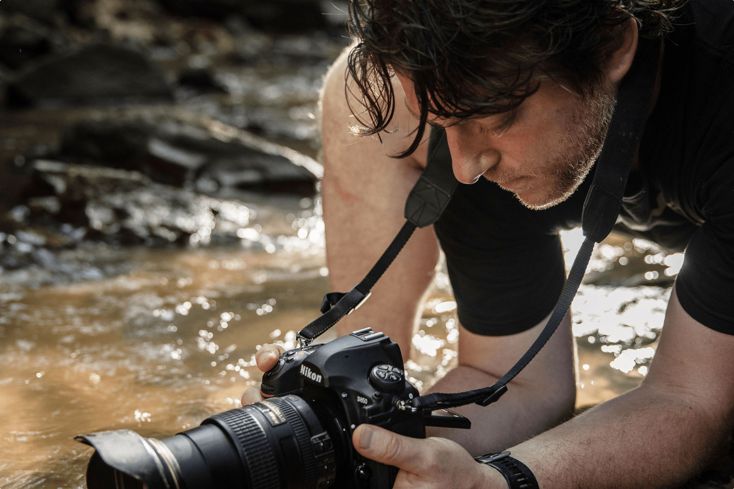 An up-close of a photographer crouching in a stream hold a camera above the water stream, positioning to take a photo