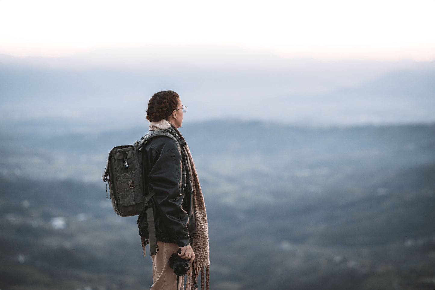 A women holding a camera equipment, and wearing a backpack and a out-of-focus mountain scenery