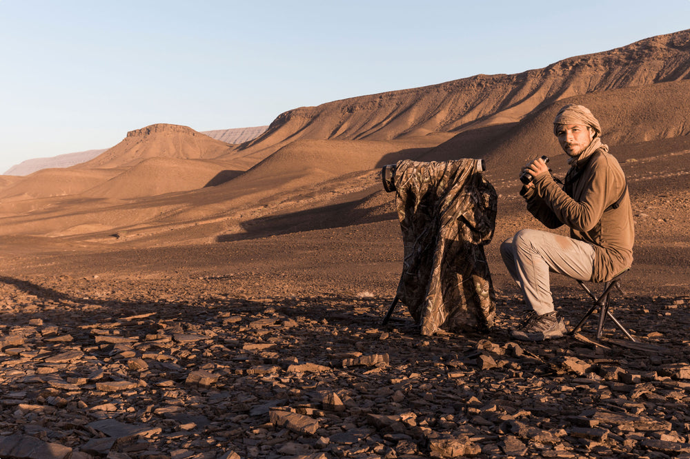 A photographer sitting holding binoculars, in front of him a camouflage tarp covering the camera with long lens 