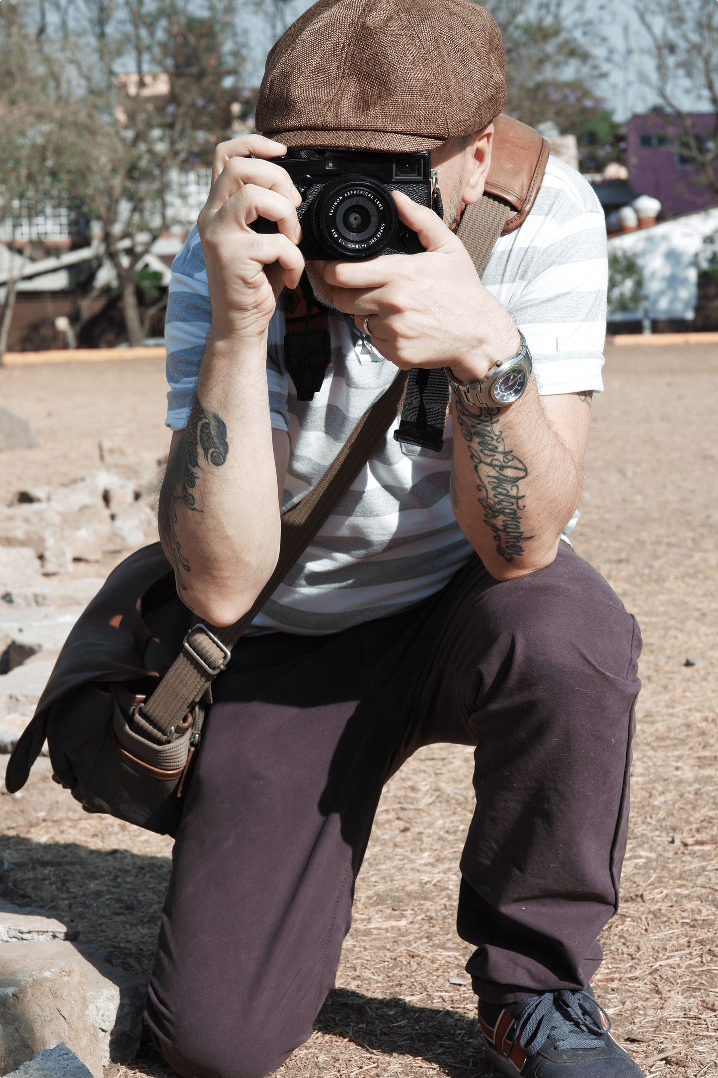 A photographer crouching down facing you, and holding a camera and taking your photo, wearing a messenger bag