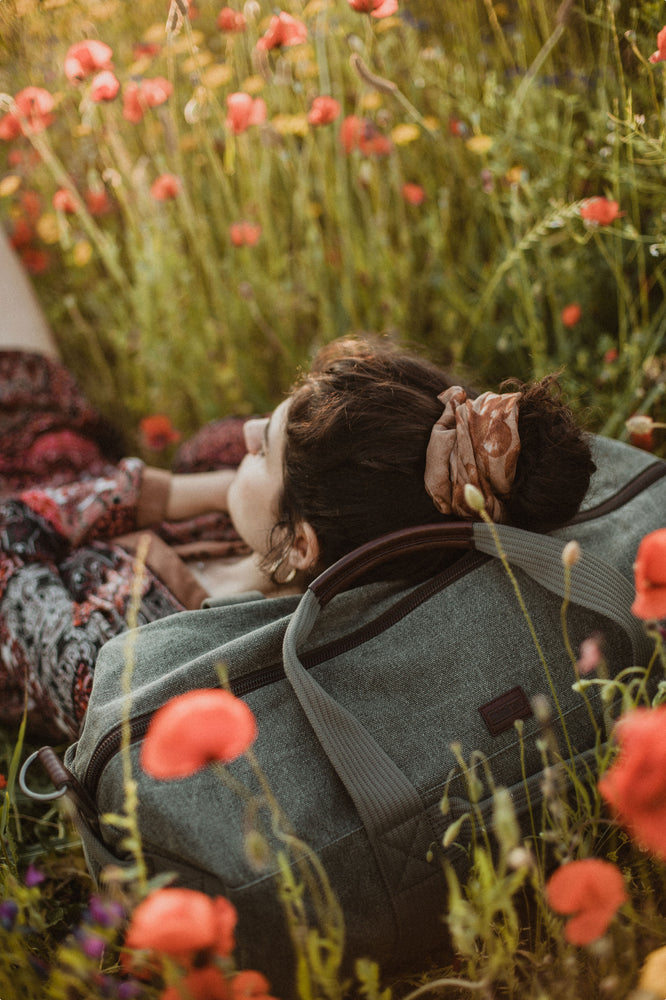 A women lying down in a field of flowers, resting her head on a duffle bag