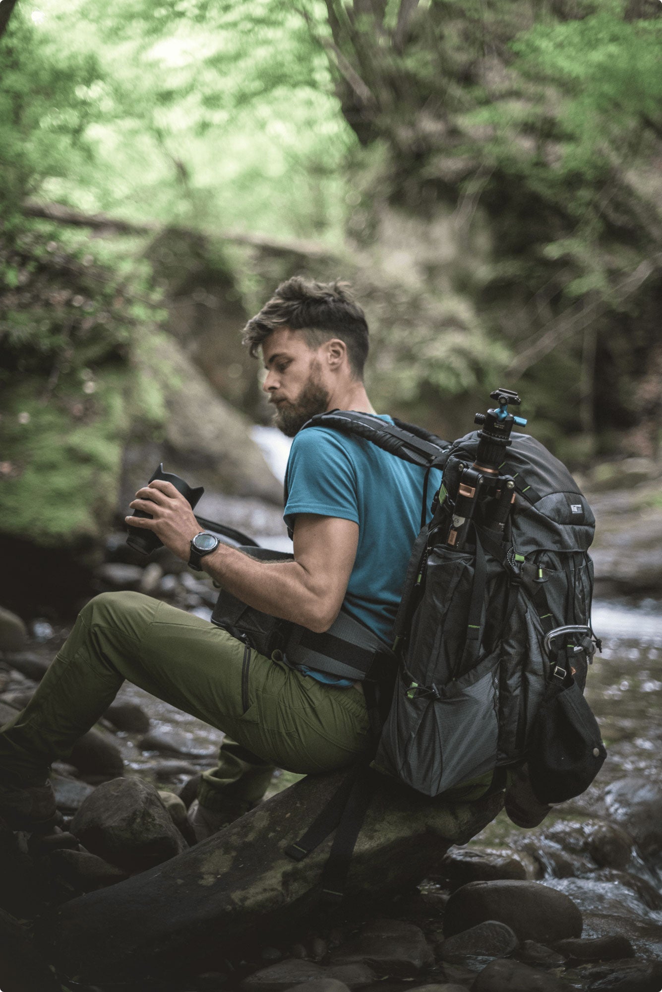 A photographer sitting on a rock wearing a backpack, and looking at his camera equipment 
