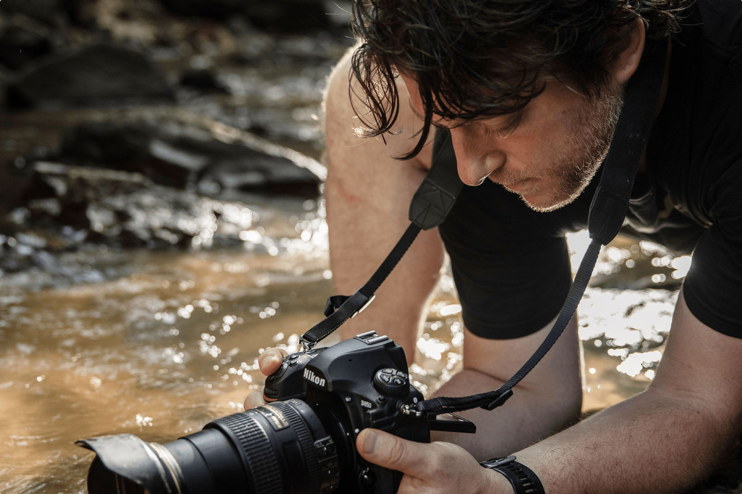 An up-close of a photographer crouching in a stream hold a camera above the water stream, positioning to take a photo 