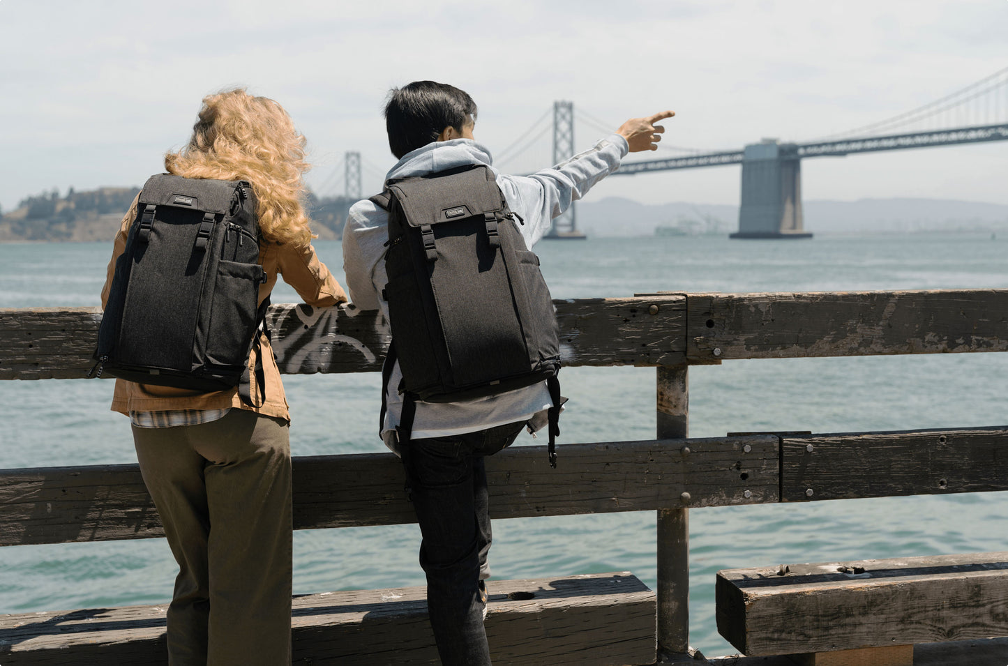 two people wearing a backpack, leaning on a fence overlooking a bridge