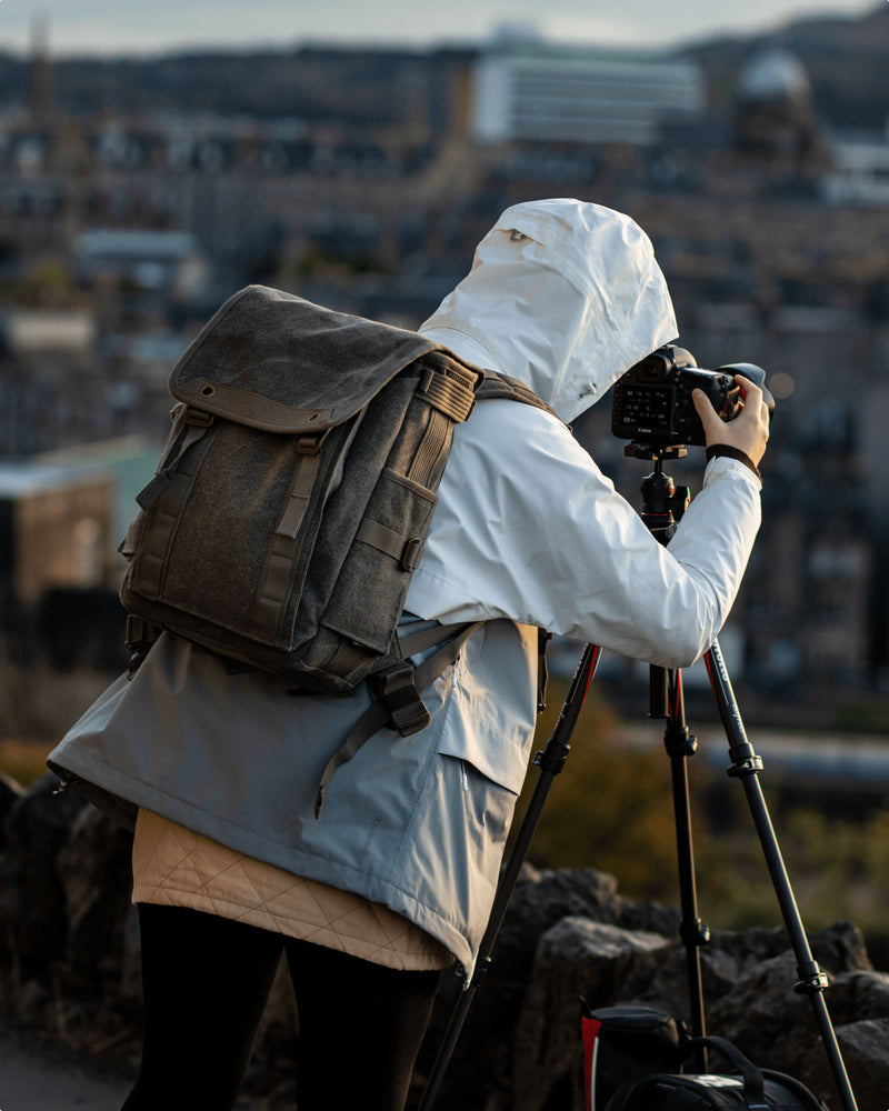 A photographer standing, wearing a backpack in front of a camera on a tripod.
